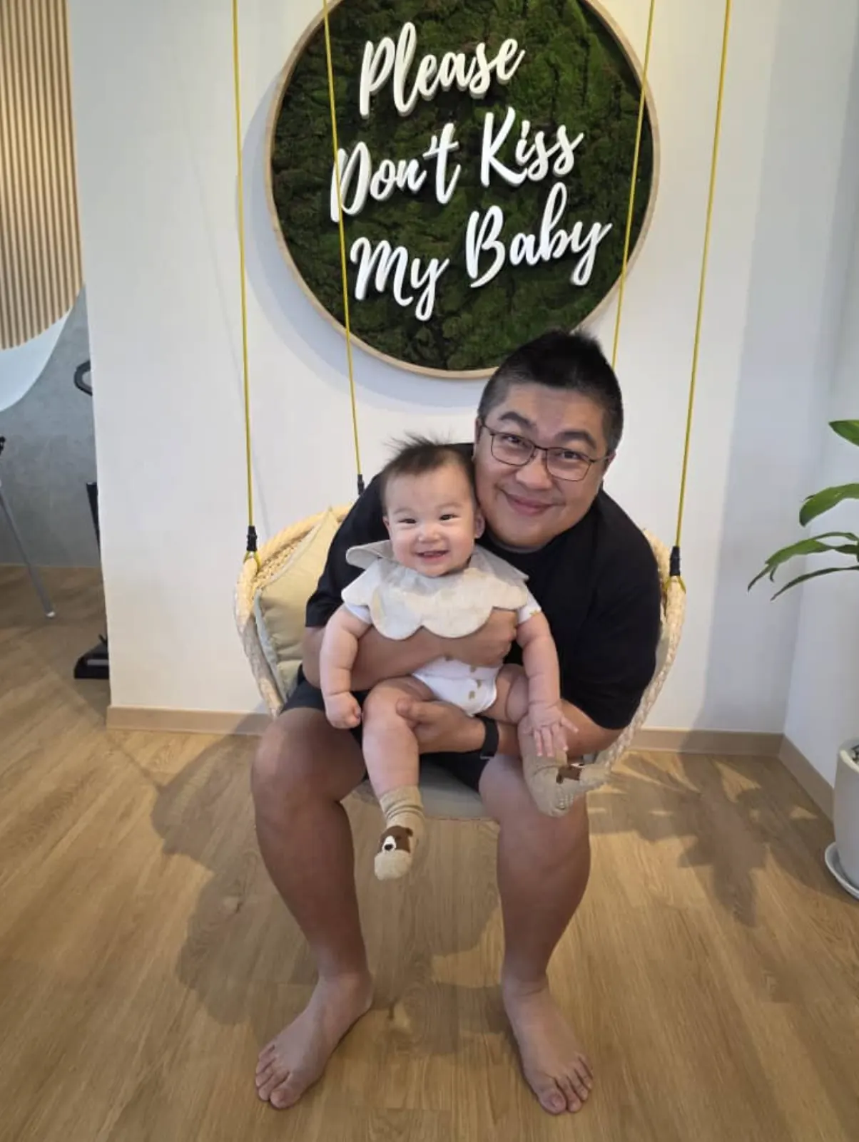 A dad holding his baby on the swing chair beneath the 'Please Don't Kiss My Baby' sign at Naru.