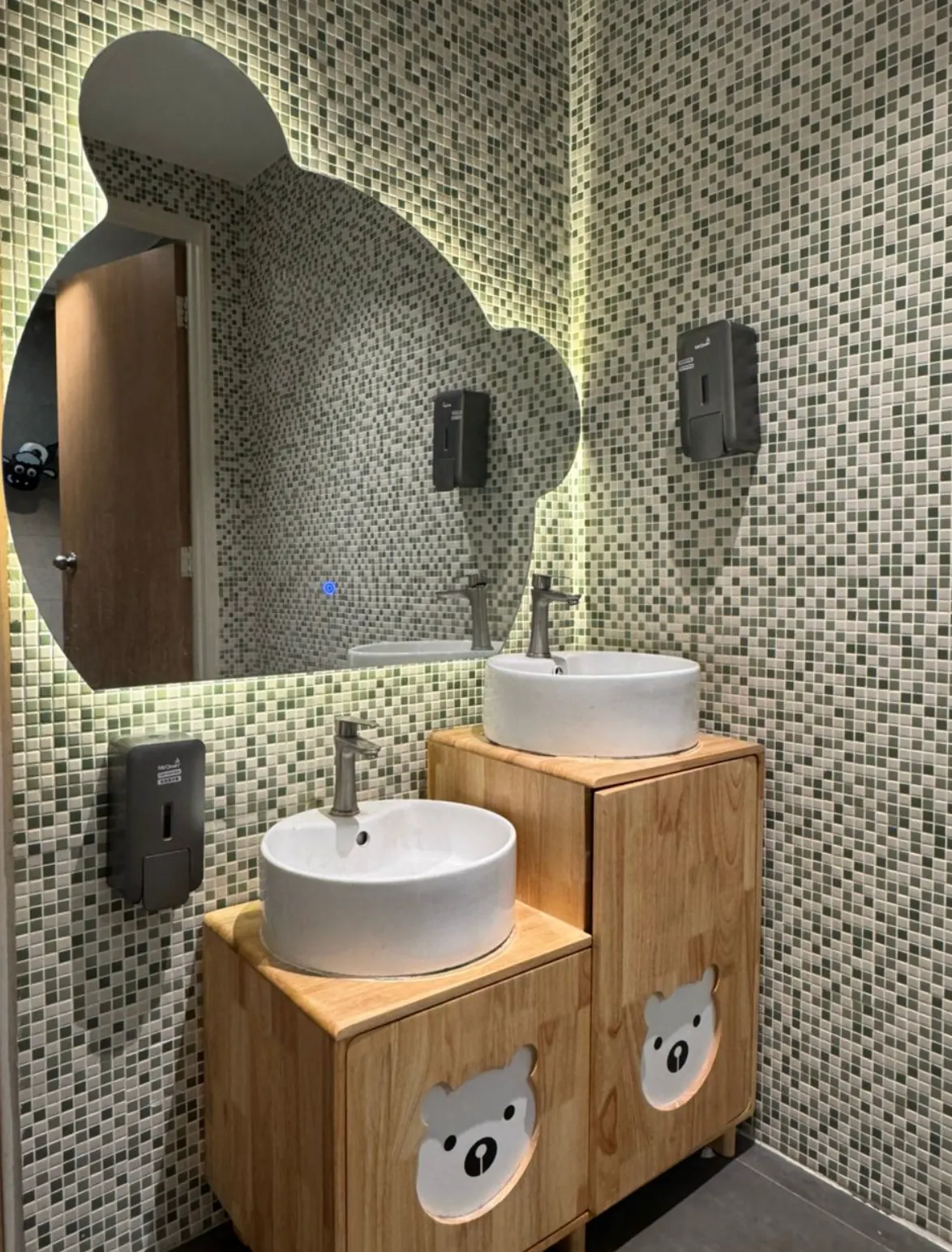 Two basins at different heights with bear-shaped cabinet fronts and a bear-shaped mirror in Naru's mosaic-tiled bathroom.