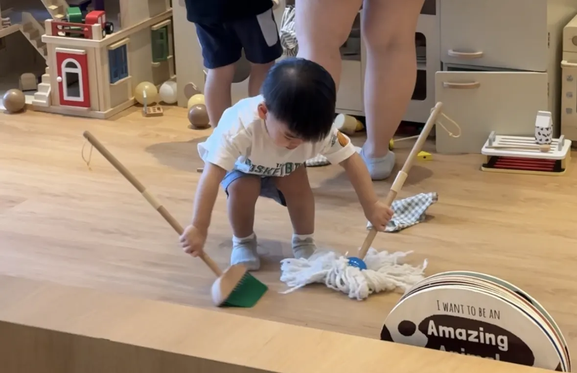 A toddler at Naru holding a broom almost as tall as he is, sweeping the floor.